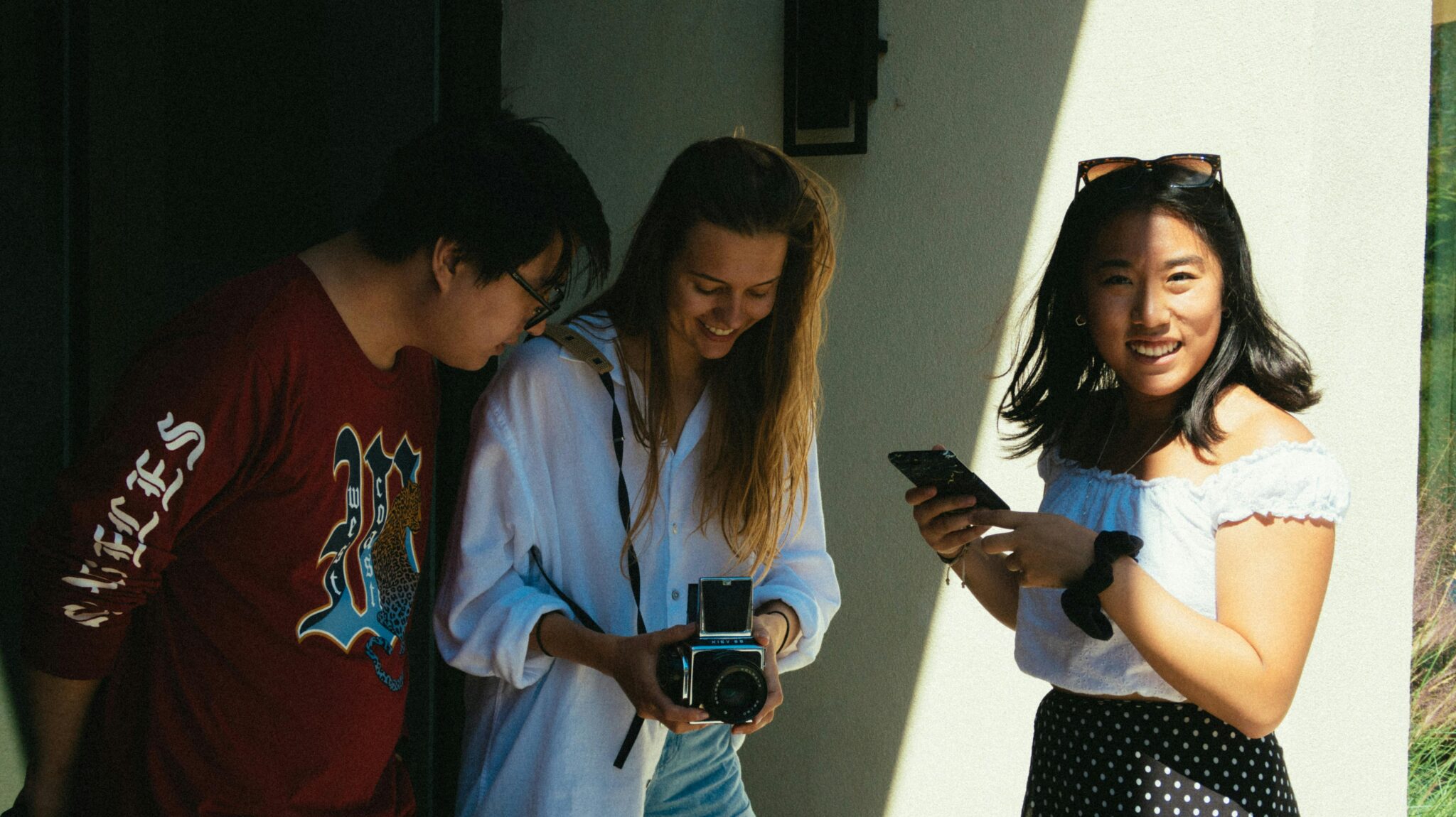 Three young friends smiling, sharing moments with a camera and phone outdoors, enjoying a sunny day.